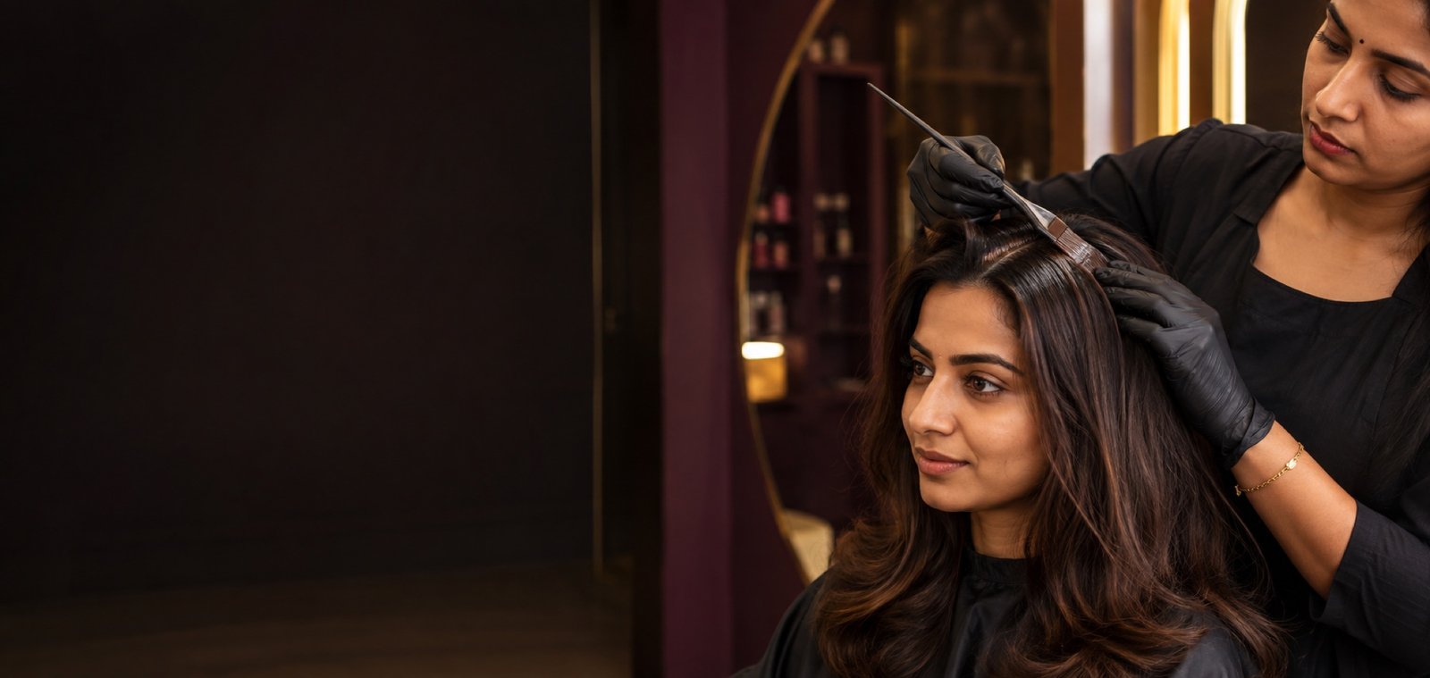 Indian colourist applying a precise dark-brown root touch-up along an Indian client's parting